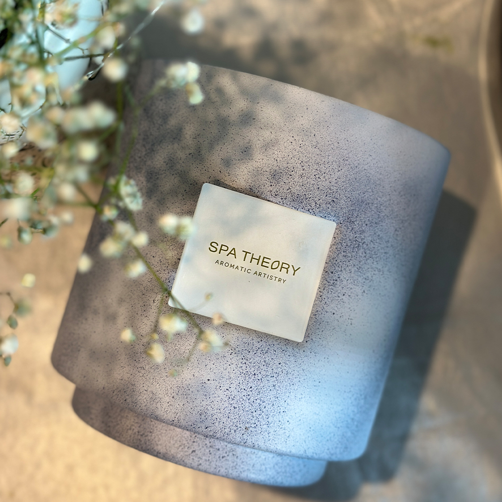 A scented candle with a label that reads 'Spa Theory - Aroma Therapy', placed on a surface with a background of flowers.