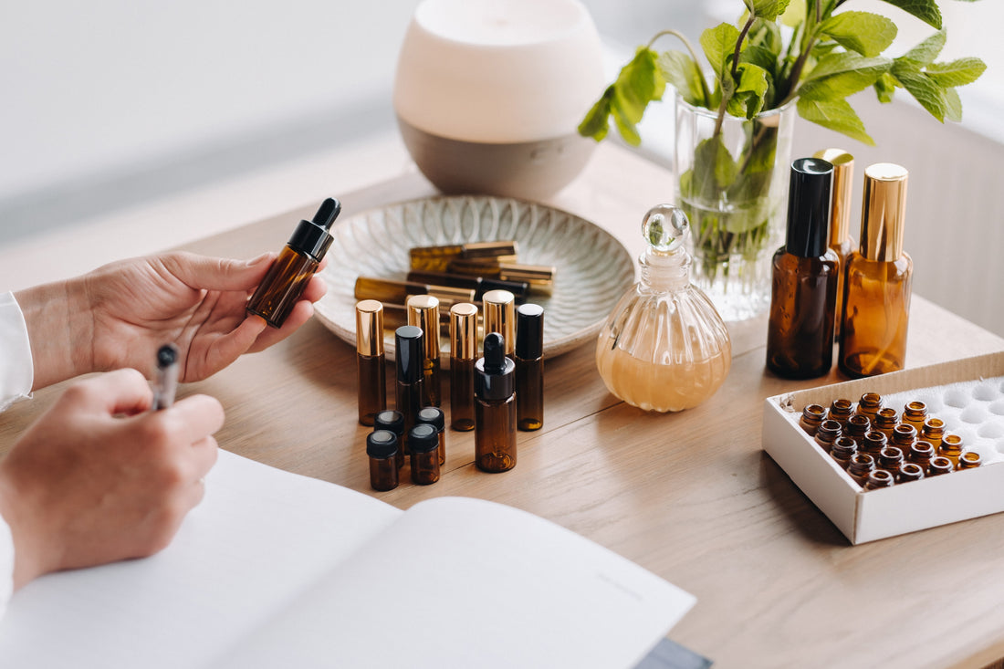 Hands take notes next to amber glass bottles, a dropper, and a potted plant on a wooden table, suggesting a serene, aromatic workspace.