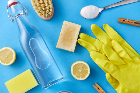 Cleaning essentials on a blue background include a glass bottle, yellow gloves, sponge, lemon halves, soap, brush, baking soda spoon, and clothespins.